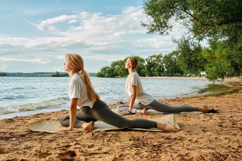Two women engage in yoga poses on the sandy beach beside a calm lake. The sun shines brightly,