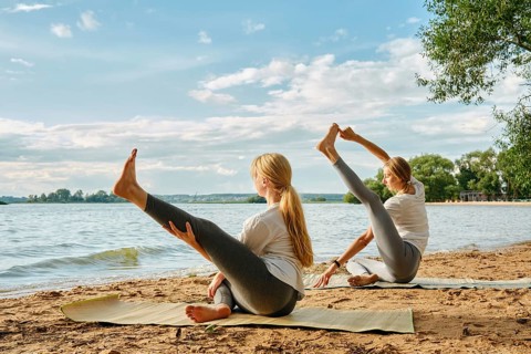 Two women practice yoga on mats by the lake, enjoying the sunny afternoon and calm waters. They