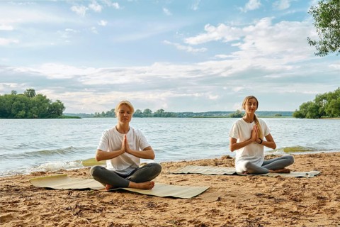 Two women are sitting cross-legged on yoga mats by the riverbank, practicing yoga with serene