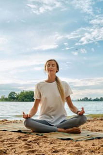 A woman sits cross-legged on a mat by the lake, practicing yoga in a serene environment. The sun