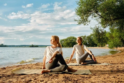 Two women practice yoga on a sandy lakeshore, surrounded by the calm waters and lush greenery. The