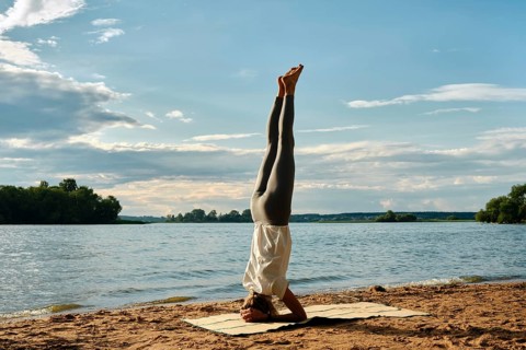 A woman performs a headstand yoga pose on a sandy beach beside a tranquil river. The sun sets in