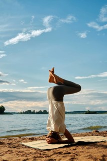 A slender woman performs a headstand yoga pose on a mat in the sand near a tranquil lake. The