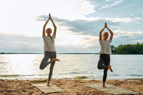Two women perform yoga poses on mats at the beach during sunset. They are surrounded by a tranquil