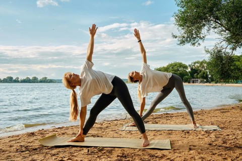 Two women practice yoga poses on the sandy shore of a lake during sunset. The sun casts a warm glow