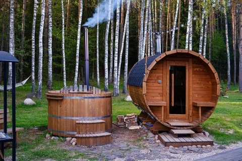 A wooden sauna and a hot tub are nestled in a lush forest. The hot tube emits smoke from its