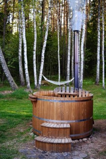 A rustic wooden hot tub sits in a peaceful clearing, surrounded by tall trees and green grass. A