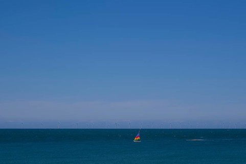 Sailboat, colourful sail, sailing on sea, sky, blue, behind offshore wind farm, wind turbines,