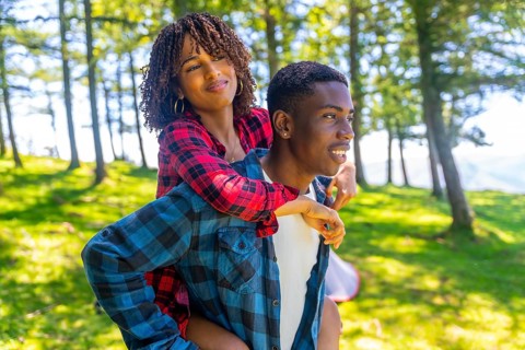 Young man giving piggyback ride to his girlfriend in forest during camping trip, enjoying summer
