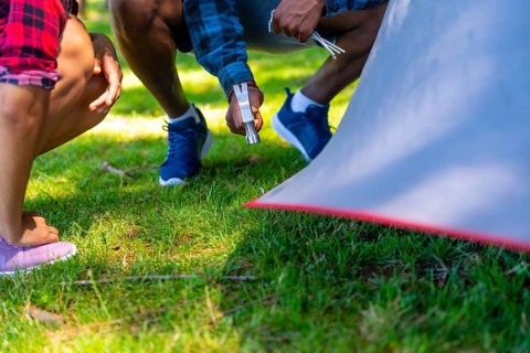 Campers are setting up their tent on a grassy campsite, using a hammer to secure the pegs and