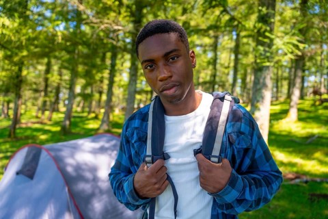 Young black man adjusts his backpack straps while standing in a forest clearing near his tent,