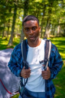 Young black man is wearing a backpack and standing in a forest near his tent, enjoying the peace of