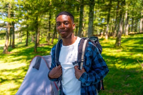 Young black man wearing a backpack, enjoying a camping trip in the woods, surrounded by lush trees