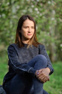 A woman sits quietly in an outdoor area, surrounded by lush greenery and soft light. She appears