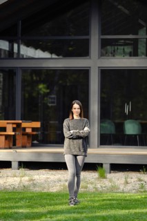 A young woman stands crossing her hands on the lawn in front of her modern house with large glass