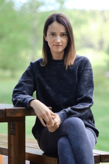 A woman is seated on a wooden bench, dressed in a black sweatshirt and leggings. She gazes directly