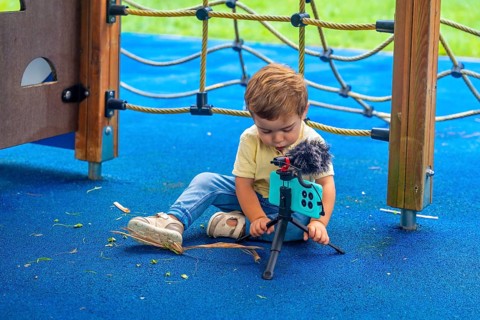 Little child sitting on playground's rubberized floor setting up a smartphone on a tripod with a