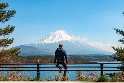 Young man sitting on railings next to a road and looking across the lake to Mt Fuji volcano, Motosu