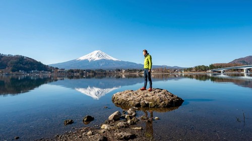 Young man standing on a rock in water, volcano Mt. Fuji is reflected in Lake Kawaguchi, Yamanashi