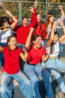 Young women sitting on stadium bleachers, cheering loudly with raised arms and painted faces,