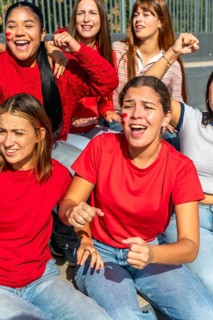Group of enthusiastic young women friends wearing red shirts and face paint, actively cheering and