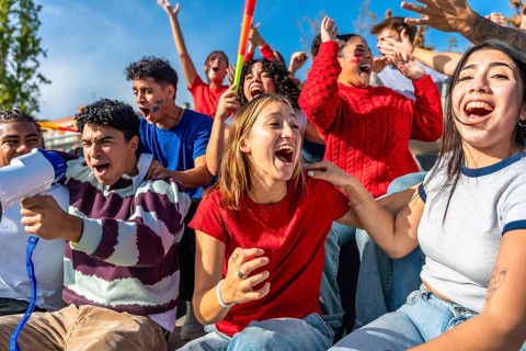Diverse group of young adult sports fans cheering and shouting with arms raised in stadium stands,