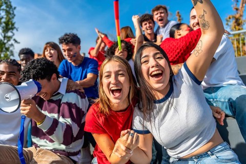 Group of excited young people watching a sports game and cheering with hands up and megaphones,