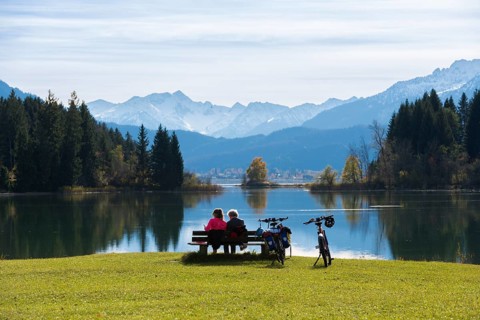 Two woman sitting on a bench look at the peaceful Forggensee with mountain views, surrounded by