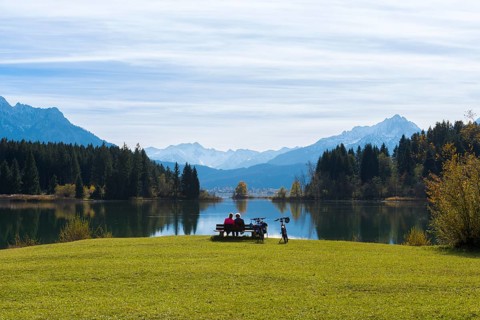 Two woman sitting on a bench at Lake Forggensee with a view of mountains, surrounded by autumn