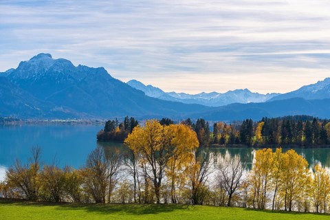 View of quiet Forggensee with mountains in the background, autumn trees on the shore, near