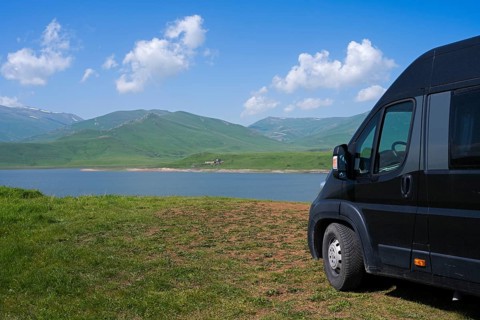 A black van stands at a lake with mountains in the background under a blue sky with clouds, camper,