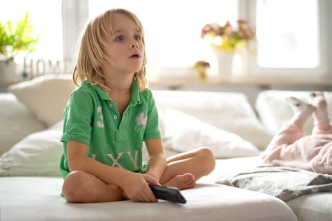 Child, boy, 7 years old, blonde, sitting on sofa, with remote control in hand, watching TV,