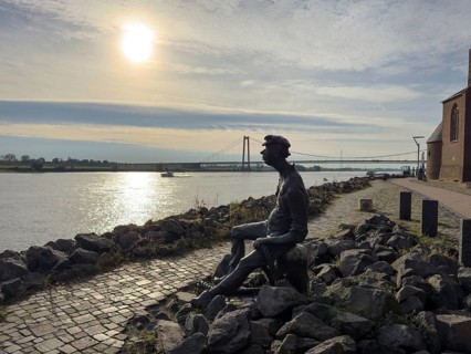 Photo with melancholy mood of bronze sculpture Poortekerl looking at Rhine waiting for ship to