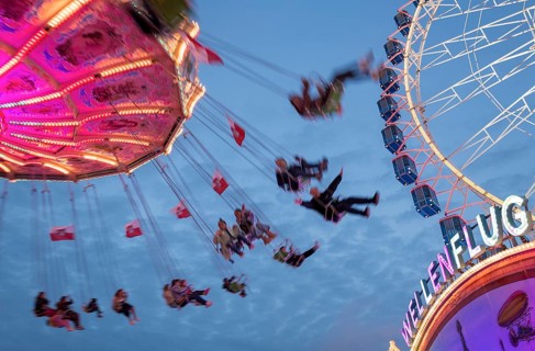 An illuminated chain carousel turns at night at a funfair, with floating people and colourful
