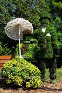 Live plant sculpture of Victorian man standing and woman with sun shade umbrella sitting on wooden
