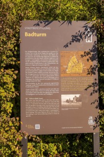 Information board about the Badturm with historical text and map, surrounded by green plants,