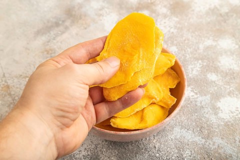 Dried Mango in ceramic bowl with hand on brown concrete background. Side view, close up. healthy