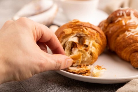 Croissant on white plate with hand on brown concrete background and linen textile, cup of coffee,