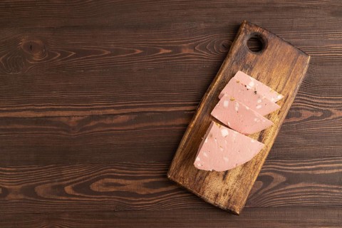 Mortadella sausage on wooden cutting board with pepper and herbs on brown wooden background. Top