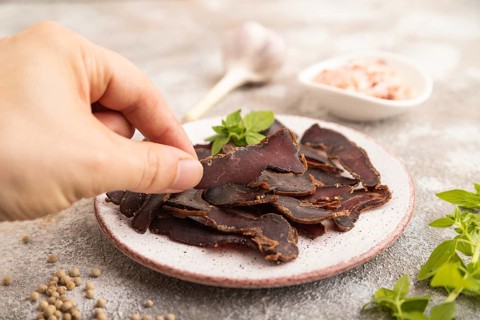 Armenian Basturma dried meat with hand on plate with pepper and herbs on brown concrete background.