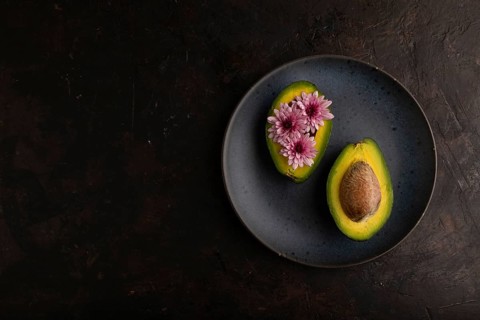 Two Avocado halves with pit and Chrysanthemums on blue plate on black concrete background, top