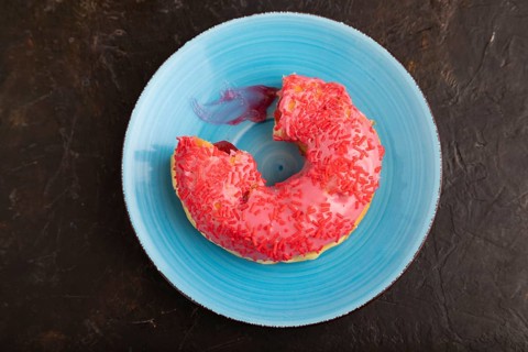 Bitten Pink Donut with sprinkles on blue ceramic plate on black concrete background, top view, flat