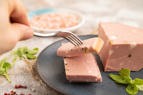 Mortadella sausage with hand on plate with pepper and herbs on brown concrete background and linen