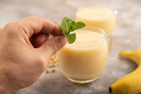 Organic non dairy banana and soy milk in glass with hand on brown concrete background. Vegan