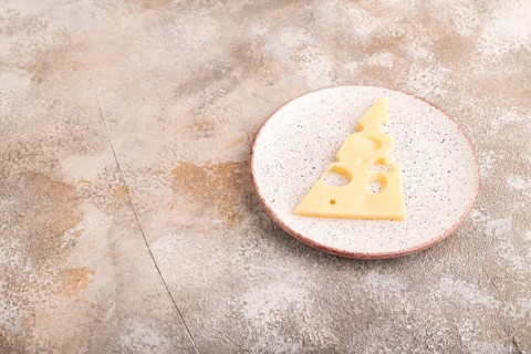 Piece of cheese with holes on ceramic plate on brown concrete background, side view, copy space,