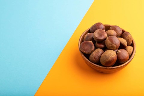 Wooden bowl with raw edible ?hestnuts on blue and orange paper pastel background, side view, copy