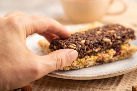 Granola bars with caramel, nuts, flakes in ceramic plate on brown concrete background with hand,