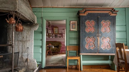 Raum mit großem alten Kamin, einem bemalten Bauernschrank und grüner Holzwand, historischer