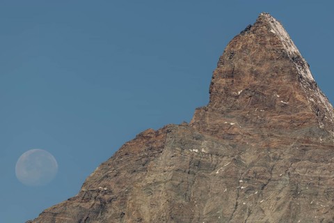 The Matterhorn mountain peak rises steeply against a bright blue sky. The full moon glistens gently