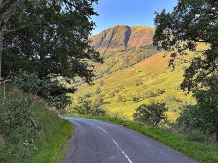 A winding road leads through a green landscape under a clear sky with a mountain in the background,